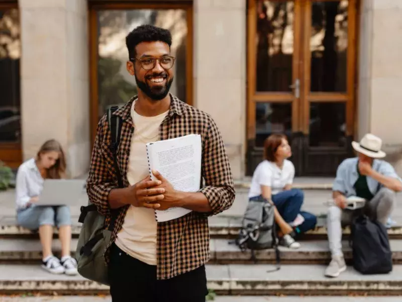 Education -Happy young university student with book standing outdoors in front of campus